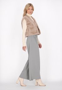 Beige Kunstfellweste über einem cremefarbenen Rollkragenpullover, kombiniert mit grauen, lockeren Hosen und beigen klobigen Stiefeln mit Absatz. Glatte Texturen und geschichtetes Design.