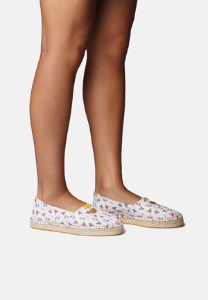 Nogi w białych wsuwanych espadrylach z kolorowymi kwiatowymi wzorami i plecionymi podeszwami z juty, przedstawione na jednolitym białym tle.