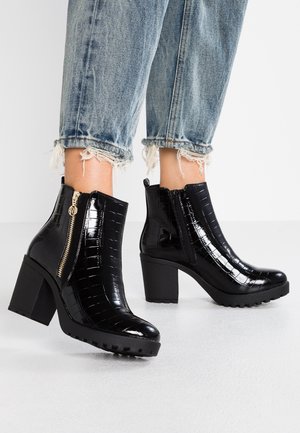 Černé lakované kozačky do kotníků s krokodýlím vzorem, blokový podpatek, dekorativní zipy a zpevněná podrážka. Na kotnících roztrhané džíny.