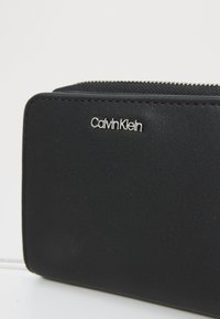 Fekete cipzáros pénztárca sima műbőrből, elöl ezüst színű "Calvin Klein" logóval. Téglalap alakú, textúrált kiegészítőkkel.