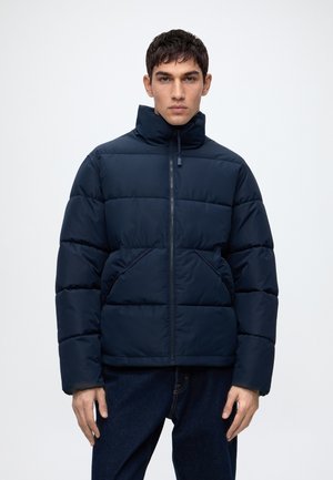 Homme portant une veste matelassée bleu marine avec un col montant et des poches avant, debout devant un fond clair uni.