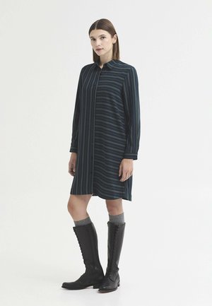 Femme portant une robe chemise rayée bleu marine associée à des chaussettes grises et des bottes noires hautes à lacets, debout devant un fond uni.