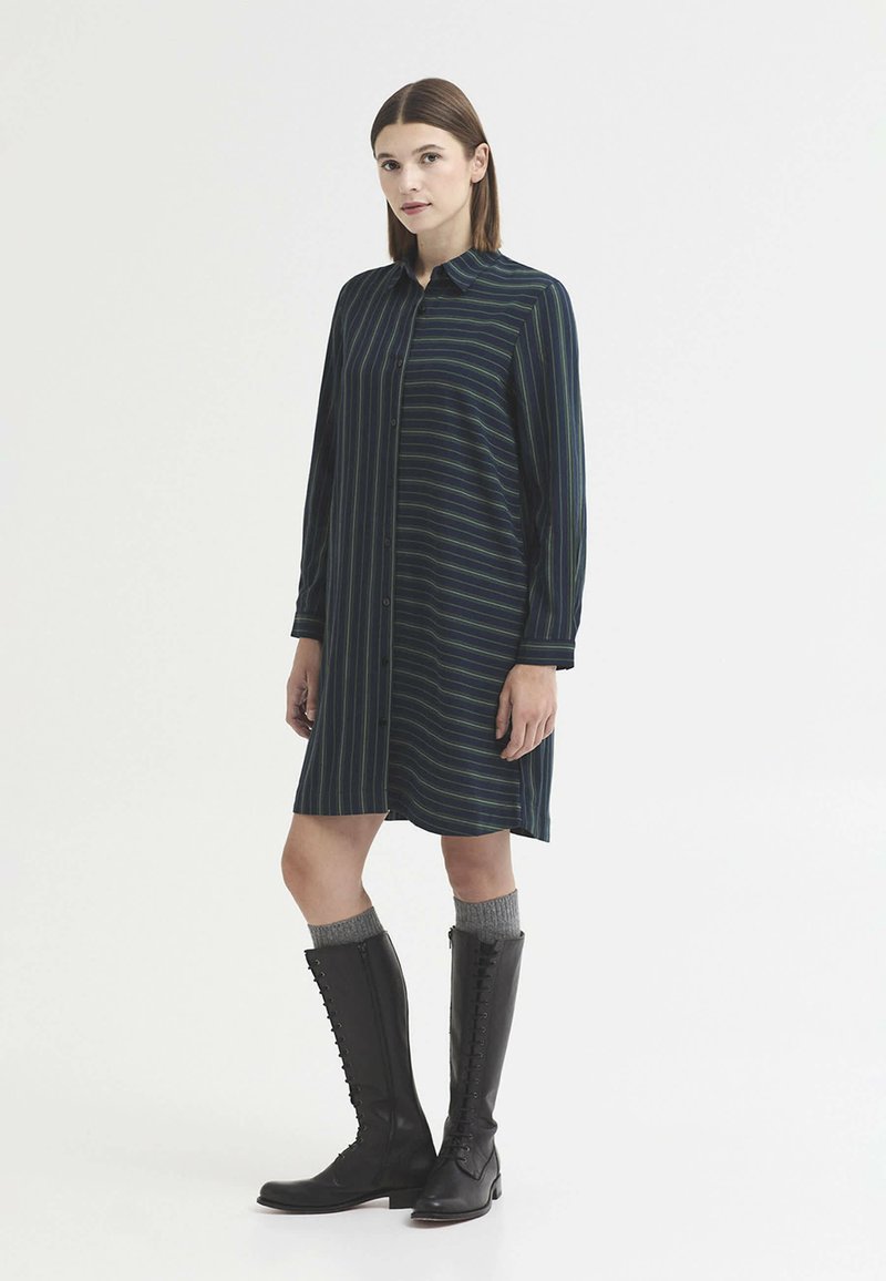 Femme portant une robe chemise rayée bleu marine associée à des chaussettes grises et des bottes noires hautes à lacets, debout devant un fond uni.
