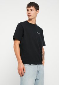 Sort bomulds T-shirt med korte ærmer, rund hals og et lille hvidt broderet logo på brystet. Sammen med lyseblå denimjeans.