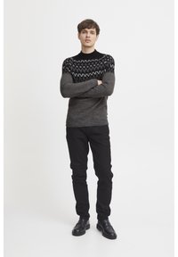 Grauer Strickpullover mit schwarzem gemustertem Schulterbereich, langen Ärmeln und Rundhalsausschnitt. Kombiniert mit schwarzen Hosen und schwarzen Schuhen, vor einem schlichten Hintergrund stehend.