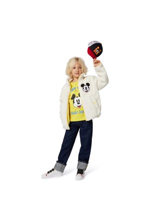 Hvid fleecejakke med Mickey Mouse-patch, gul grafisk t-shirt, mørkeblå jeans og hvide sneakers med sorte og røde detaljer.