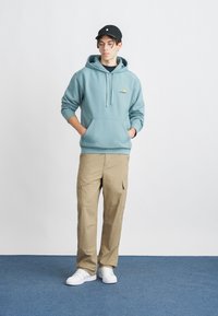 Hanorac cu glugă de culoare albastru deschis, cu buzunar frontal, pantaloni cargo khaki și adidași albi. O șapcă neagră cu logo completează ținuta.