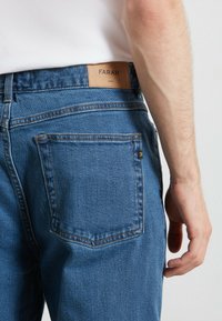 Jeans en denim bleu avec une étiquette en cuir à la taille portant le logo "FARAH" en relief. La poche arrière est simple avec une petite étiquette logo.