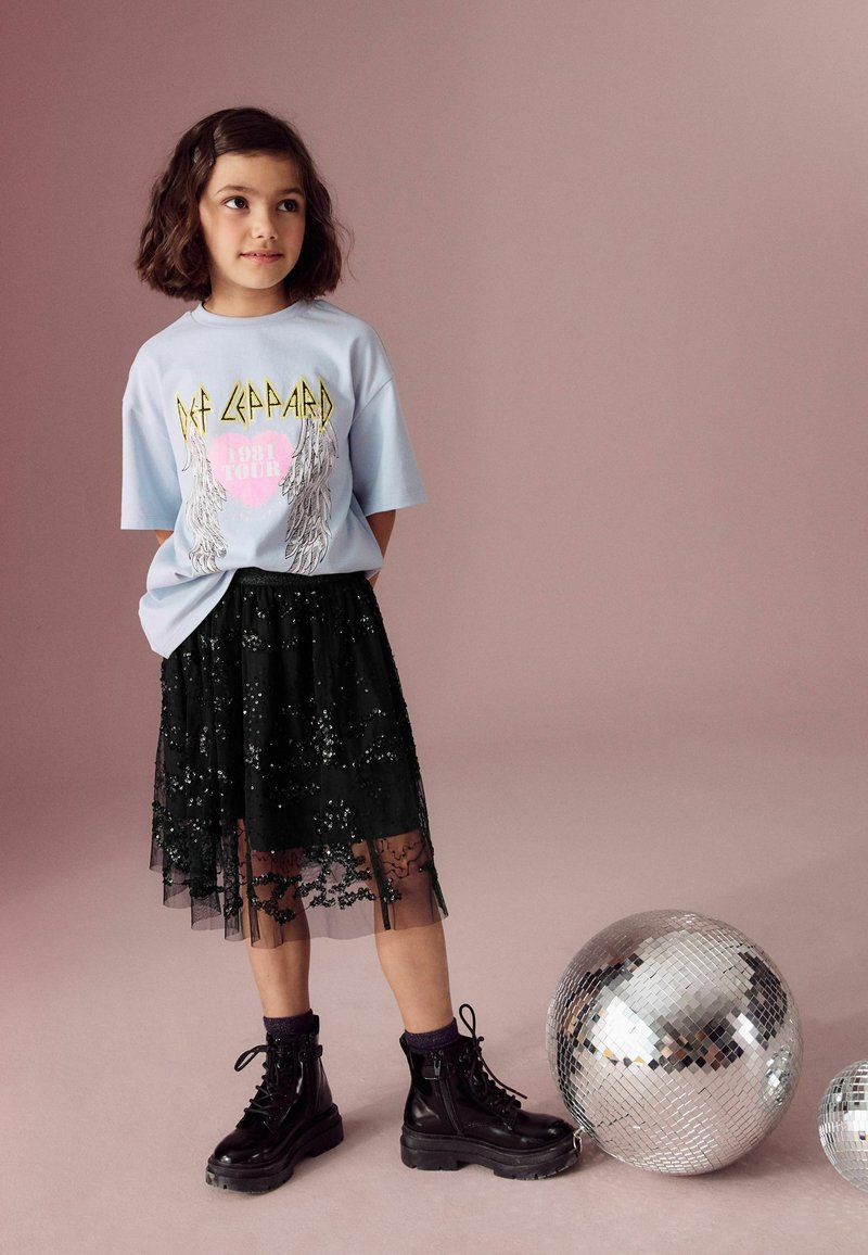 Lichtblauwe graphic t-shirt met de tekst "Def Leppard", gecombineerd met een zwarte, paillettengevoerde tule rok en zwarte veterschoenen. Twee discoballen in de buurt.