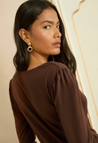 Brązowa bluzka z bufiastymi, długimi rękawami. Modelka nosi duże, okrągłe złote kolczyki podkreślające strój. Gładka tkanina o subtelnym połysku.