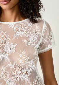 Witte kanten top met bloemenpatronen, korte mouwen en gegolfde randen. Doorzichtige, delicate textuur met intrigerende details. 