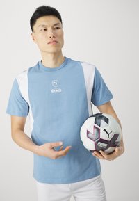 Šviesiai mėlyna marškinė su baltomis šoninėmis juostelėmis, centre yra logotipas. Modelis laiko juodai baltą futbolo kamuolį su spalvotais akcentais.