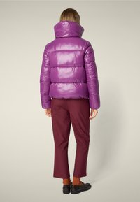 Shiny purple puffer jacket with high collar and prominent quilted design, paired with dark burgundy trousers and black shoes.