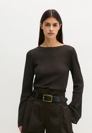 Kobieta o długich ciemnych włosach, ubrana w ciemny prążkowany top z długim rękawem, spodnie z wysokim stanem i czarny pasek ze złotą klamrą.