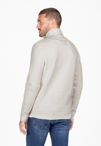 Jersey de color crema con cuello alto, diseño de punto texturizado y puños ajustados, combinado con jeans azules. Se muestra la vista trasera.