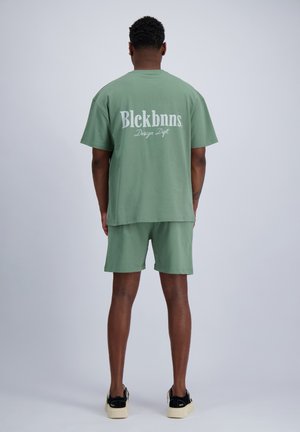 Man met zijn rug naar de kijker toe, draagt saliegroene korte broek en bijpassend oversized T-shirt met de tekst "Blckbnns Design Dept." op de achterkant, zwarte en beige sneakers.