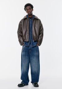 Hned�á umelá kožená bunda s kapucňou na modrej rifľovej bunde, v kombinácii s širokými modrými rifľovými nohavicami a čiernymi masívnymi topánkami. Viditeľné strieborné reťazové detaily.