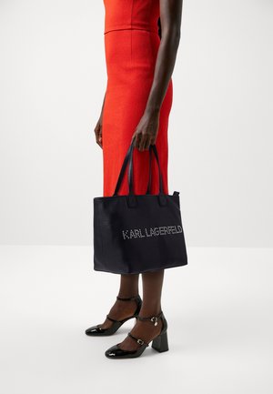 Sac fourre-tout en cuir noir avec poignées à bandoulière et détails du logo cloutés. Le sac a une forme structurée et une texture grainée.