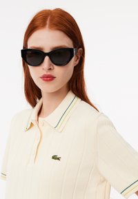 Polo shirt color crema con texture a coste e colletto a strisce verdi e gialle; include un piccolo logo ricamato a forma di coccodrillo. Occhiali da sole neri.