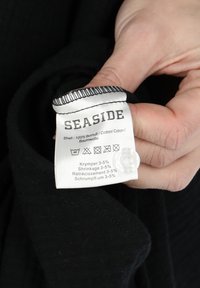 Black cotton clothing label shows the brand "SEASIDE" with care instructions and composition details. Label features a white background and black text.