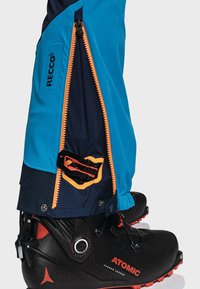 Niebiesko-granatowe spodnie narciarskie z pomarańczowym suwakiem, z logo RECCO i kieszenią na zatrzask, noszone na czarnych butach narciarskich z czerwonymi akcentami.