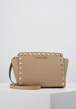 Beige Michael Kors håndledstaske med gyldne firkantede nitter langs frontkanterne og en tynd håndledsrem på en hvid overflade.