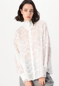 White lace blouse featuring a high collar, button front, and wavy patterns with cut-outs. Long puff sleeves with lace trim at the cuffs.