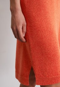 Robe tricotée orange avec un tissu texturé, une fente sur le côté et un ourlet côtelé. Gros plan d'une main contre le vêtement montrant le détail du tissage.