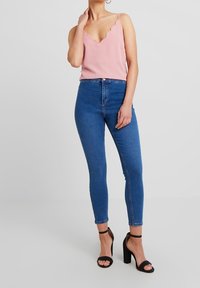 Woman wearing a pink scalloped-edge cami top, high-waisted blue skinny jeans, and black high-heeled ankle-strap sandals.