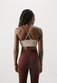 Top sportivo marrone con spalline incrociate, tessuto morbido e dettagli con logo. Abbinato a leggings bordeaux a vita alta, dalla texture elegante.