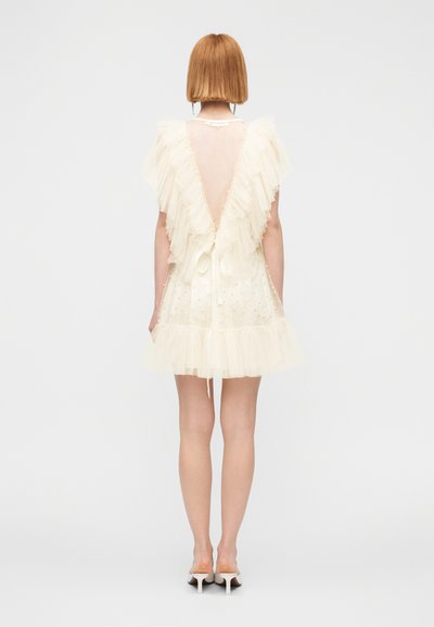 Γυναίκα με κοντά μαλλιά που φοράει φόρεμα από κρεμ τούλι με βολάν, λεπτομέρειες με πέρλες, βαθιά V πλάτη και λευκές mule γόβες με τακούνι, στέκεται μπροστά σε λευκό φόντο.