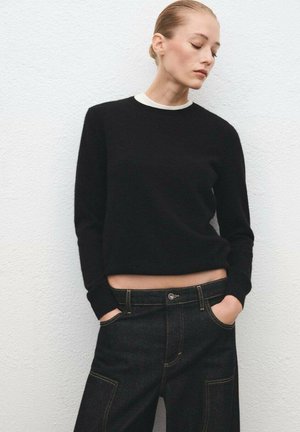 Suéter corto negro con cuello redondo, combinado con pantalones de mezclilla oscuros que presentan costuras amarillas y un corte relajado.
