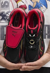 Černé a červené sportovní boty s texturovaným svrškem, zlatými akcenty a kresleným logem. Mají polstrovaný jazyk a dekorativní šněrovací smyčky.