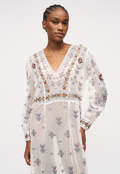 Jeune femme aux cheveux tressés portant une robe blanche transparente avec une broderie florale colorée et des manches longues, sur un fond uni.