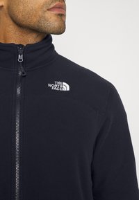 Giacca in pile con zip di colore blu scuro e colletto alto, con logo ricamato bianco sul lato sinistro del petto e dettagli in tessuto testurizzato.