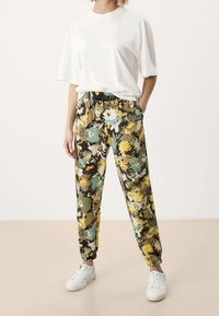 Spodnie dresowe w kwiatowy wzór w kolorach zielonym, żółtym i białym, z elastyczną talią i mankietami, zestawione z prostą, białą, oversize'ową koszulką i białymi trampkami.