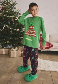 Green long-sleeve shirt features a pixelated tree and TNT graphic. Pajama pants are black with colorful Minecraft patterns. Soft green slippers.