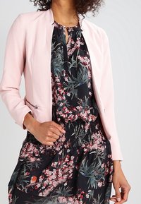 Ljust rosa strukturerad kavaj med rund hals, bärs över en blommönstrad mörk klänning med gröna och rosa accenter.