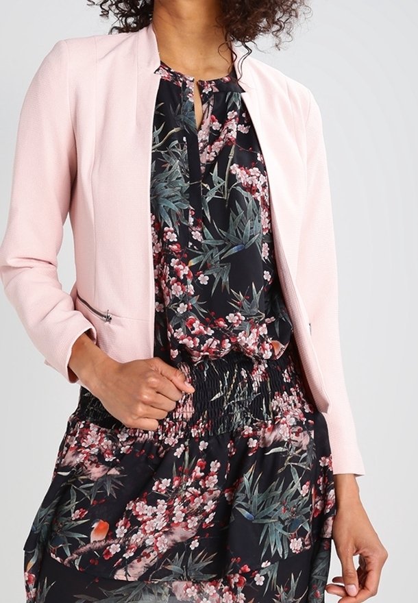 Ljust rosa strukturerad kavaj med rund hals, bärs över en blommönstrad mörk klänning med gröna och rosa accenter.