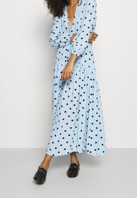 Femme portant une robe midi à pois bleue clair à manches longues avec un décolleté en V profond et des mocassins noirs, se tenant sur un sol gris.