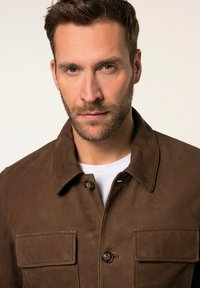 Bruine jas van textuurstof, met een kraag, knoopsluiting en twee voorzakken. Draagt over een witte shirt.