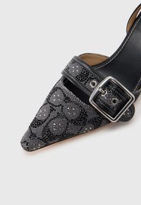 Fekete és ezüst színű hegyes orrú cipő, texturált felülettel és csillogó díszítésekkel, széles pánttal, fémes csattal.
