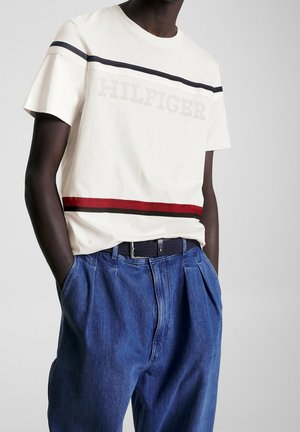 Camiseta de algodón en color crema con rayas horizontales en azul marino y rojo y el logo "HILFIGER"; combinada con pantalones de mezclilla azules sueltos y un cinturón de fibra negra.