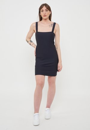 Abito nero aderente con spalline larghe, scollatura quadrata e superficie liscia, indossato con sneaker bianche su uno sfondo neutro.