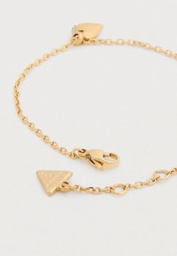 Βραχιόλι σε χρυσό χρώμα με σχεδιασμό αλυσίδας που διαθέτει τρίγωνα και καρδιές ως διακοσμητικά στοιχεία. Κούμπωμα με κλείσιμο τύπου αστακού και λείο, λαμπερό υλικό.