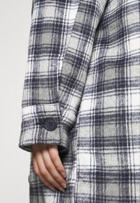 Cappotto in flanella a quadri con tonalità di grigio, bianco e blu navy. Dettagli notevoli includono un bottone sul polsino e una texture morbida.