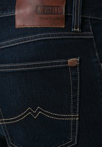 Mörkblå denimjeans med en läderetikett med "MUSTANG True Denim", synliga sömmar och en ficka med dekorativt sömdetalj.
