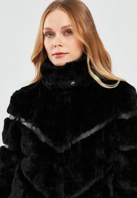 Black faux fur coat with a high collar, featuring horizontal panels and a soft, textured finish. Accented with leather details and snap closure.