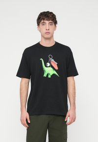 Μαύρο βαμβακερό μπλουζάκι με πράσινο γραφικό δεινόσαυρο, συνοδευόμενο από ασημένιο μπρελόκ με κόκκινο και γκρι αξεσουάρ.