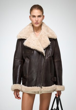 Brown shearling jacket with a large beige collar, featuring a zip closure, side pockets, and buckle accents at the hem and sleeves.